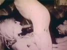 A blonde woman in black lingerie is sitting on a couch. On either side of her are two naked guys and she is jerking them both off. Then she gives one guy a blow job while the other one is fucking her from behind.